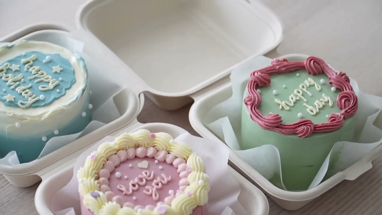 Three bento cakes in lunch boxes: a blue happy holiday cake, a sage green happy day cake and a pink love you cake