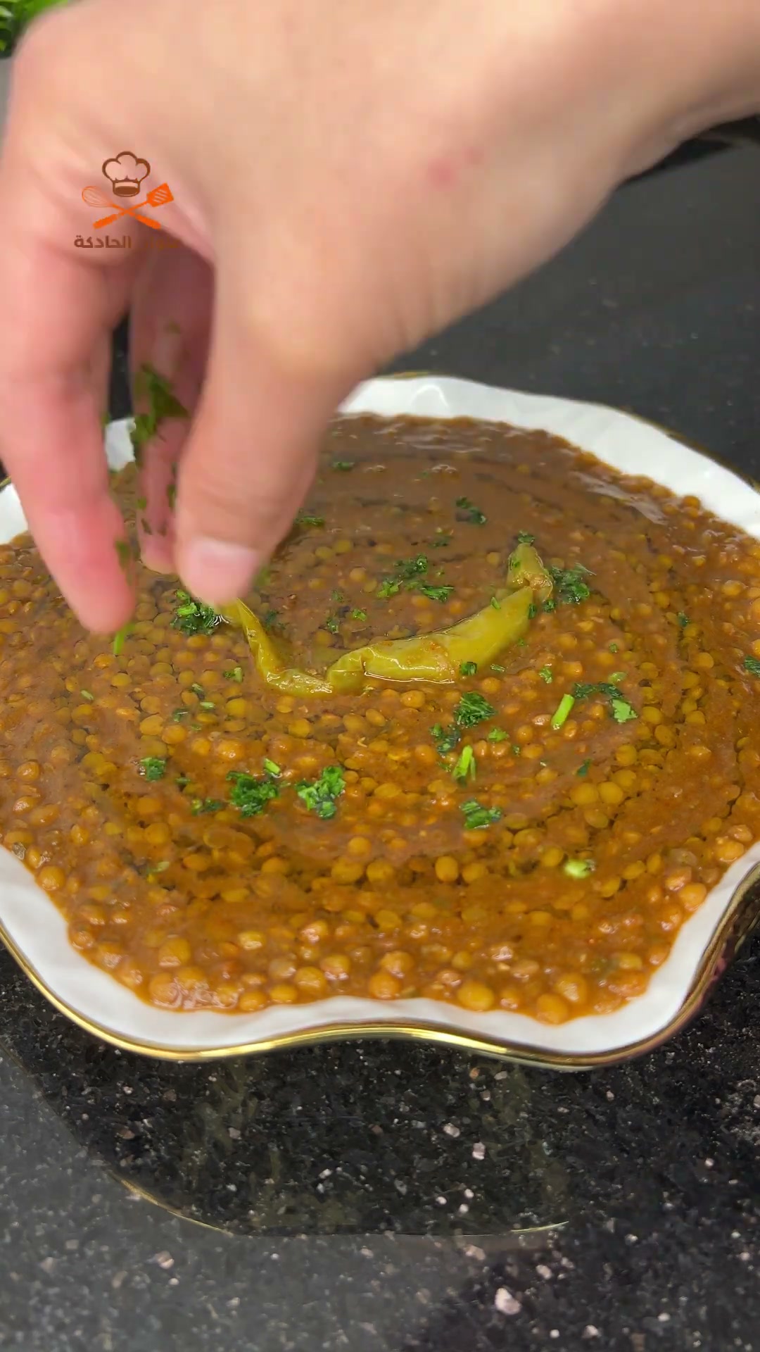 Sprinkling chopped parsley over a bowl of Moroccan lentils (l'3des) topped with a green chili pepper