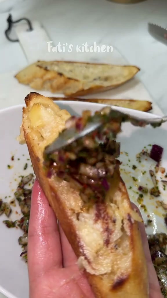 Spooning red onion relish onto a toasted baguette slice spread with roasted garlic